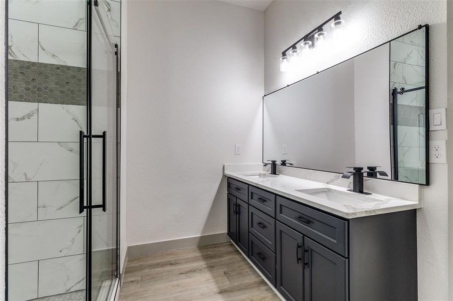 Bathroom featuring a textured wall, double vanity, a marble finish shower, and light wood-style flooring Bathroom featuring a textured wall, double vanity, a marble finish shower, and light wood-style flooring