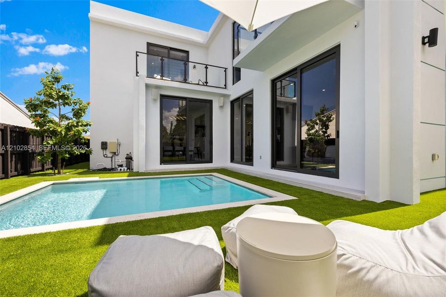 Modern exterior featuring a swimming pool, artificial turf, second-story balcony with glass railing, and floor-to-ceiling sliding glass doors