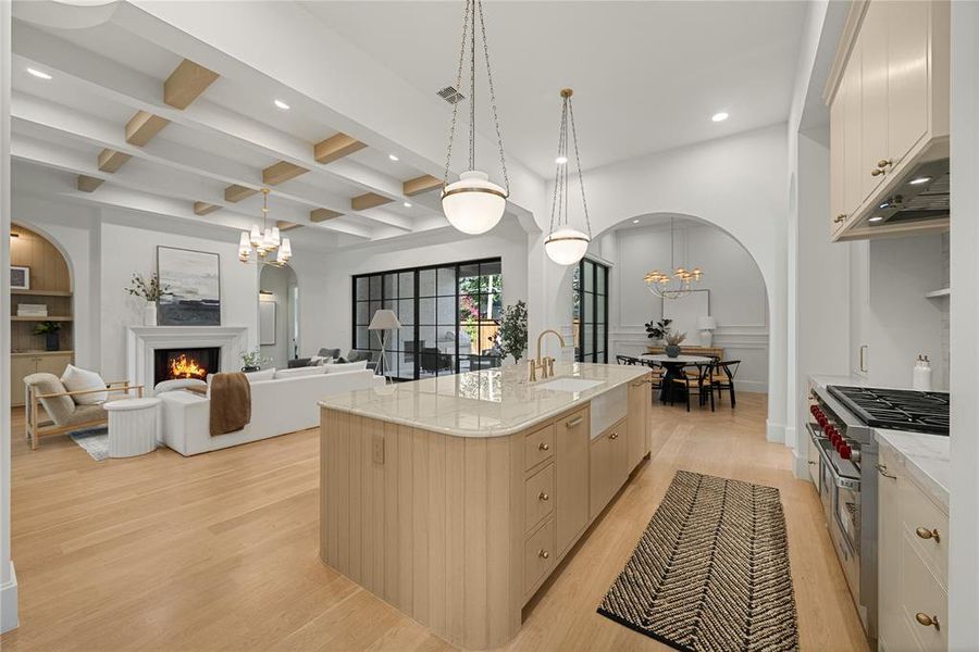 Expansive open-concept living area featuring light wood-finish flooring, a kitchen island with a farmhouse sink, a built-in gas range, a white fireplace, and exposed ceiling beams
