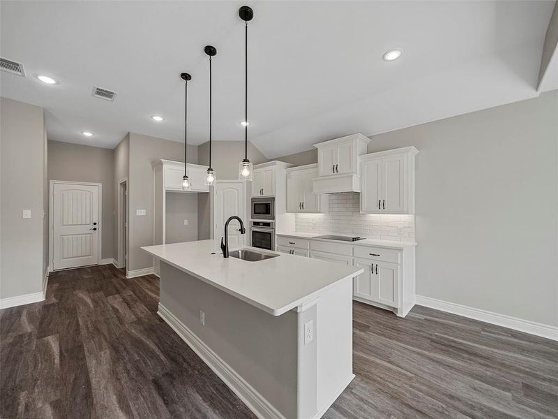 Kitchen with white cabinetry, decorative light fixtures, a center island with sink, tasteful backsplash, and dark wood-style floors Kitchen with white cabinetry, decorative light fixtures, a center island with sink, tasteful backsplash, and dark wood-style floors