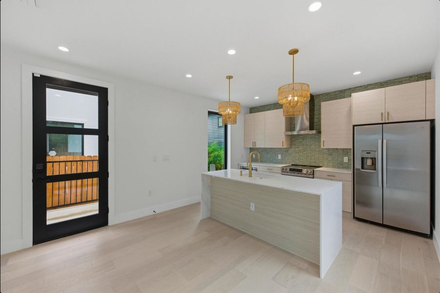 Kitchen with appliances with stainless steel finishes, light stone counters, wall chimney exhaust hood, pendant lighting, and a kitchen island with sink Kitchen with appliances with stainless steel finishes, light stone counters, wall chimney exhaust hood, pendant lighting, and a kitchen island with sink