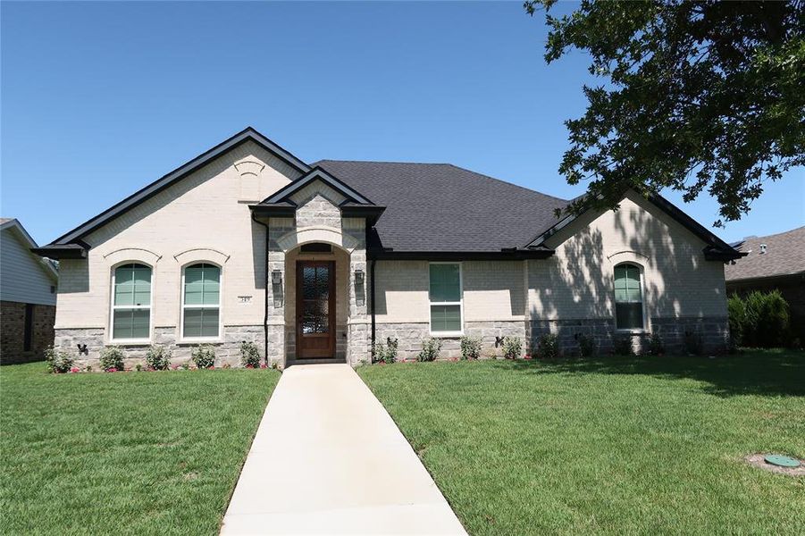 French country inspired facade featuring stone siding, a front yard, brick siding, and roof with shingles