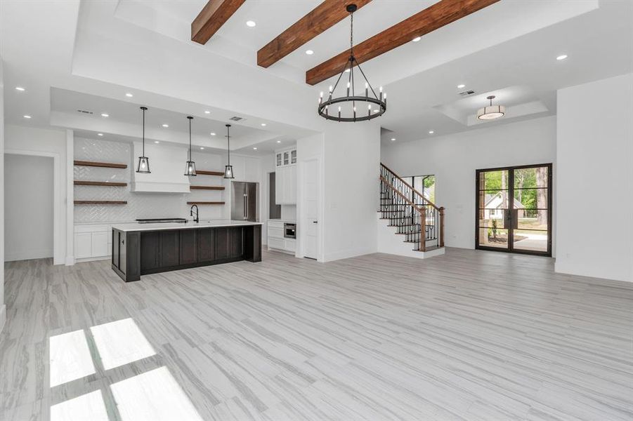 A spacious kitchen with a large island and pendant lighting, sleek white cabinetry with floating shelves, and a light-toned, wood-look flooring throughout. Large windows and glass doors provide ample natural light and a view to the outside.