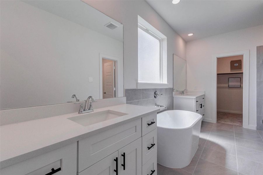 Beautiful primary bath with a long vanity, sleek countertop, large mirror, freestanding soaking tub, gray tile flooring, and a bright window for natural light. The clean white cabinetry, black hardware, and modern fixtures create a polished retreat, while the adjoining closet and shower areas add convenience and function. Beautiful primary bath with a long vanity, sleek countertop, large mirror, freestanding soaking tub, gray tile flooring, and a bright window for natural light. The clean white cabinetry, black hardware, and modern fixtures create a polished retreat, while the adjoining closet and shower areas add convenience and function.