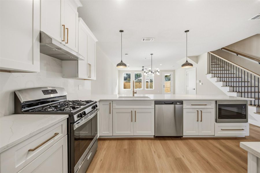 This modern kitchen features sleek white cabinetry, stainless steel appliances, and a spacious island with pendant lighting. The open layout flows into a bright dining area with large windows, and there's a stylish staircase in the background.