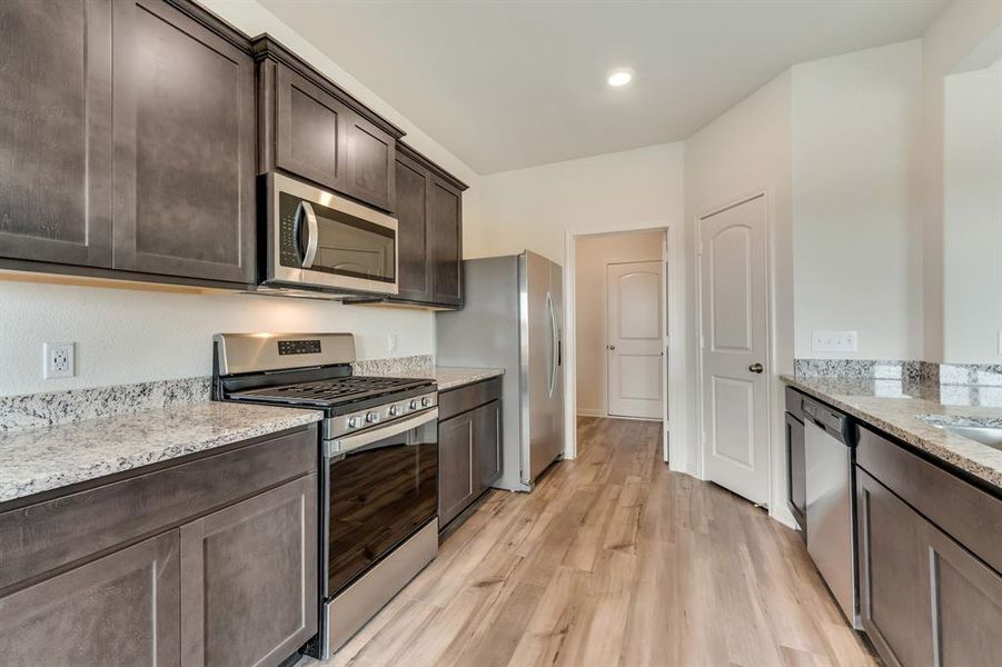 Kitchen featuring dark brown cabinetry, stainless steel appliances, light stone counters, and light wood-type flooring