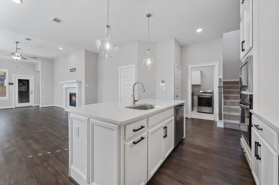 This is a spacious, modern kitchen with white cabinetry and stainless steel appliances. It features a large island, stylish pendant lighting, and opens into a bright living area with a view of the backyard through the windows and glass door.