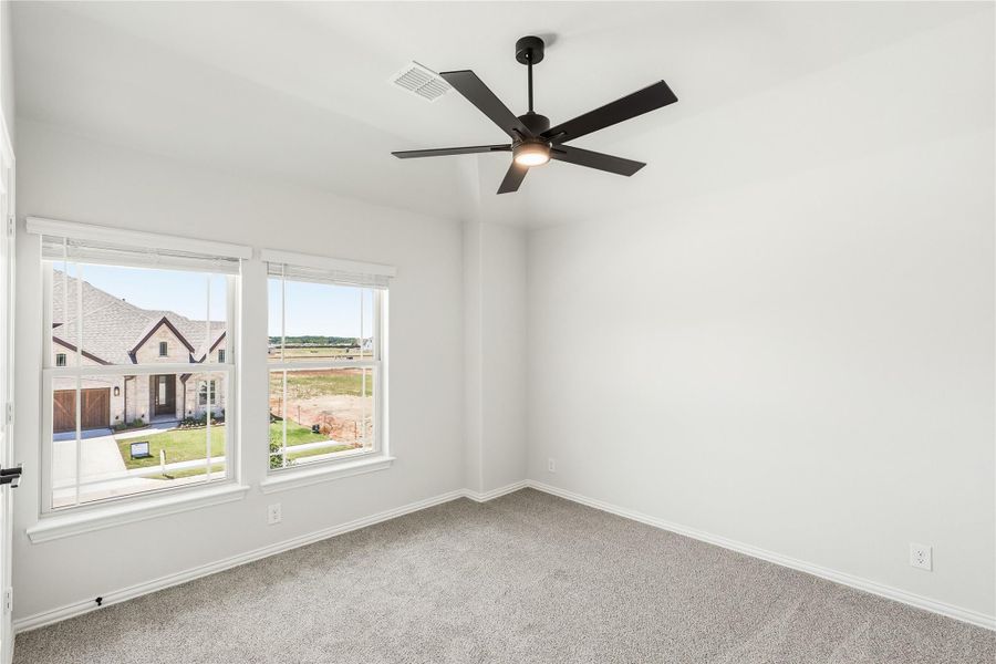 Empty bedroom with carpet flooring, black ceiling fan, and two windows overlooking neighboring homes