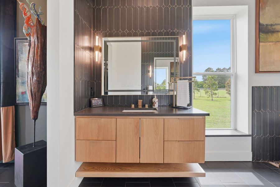 This modern bathroom features a sleek, floating wooden vanity with a dark countertop, set against stylish dark tile walls. It includes a large, well-lit mirror and a window offering a scenic outdoor view.