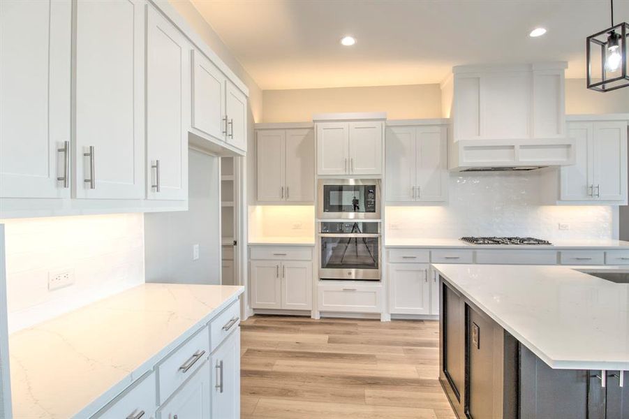 Kitchen with light wood-type flooring, backsplash, stainless steel appliances, white cabinetry, and hanging light fixtures