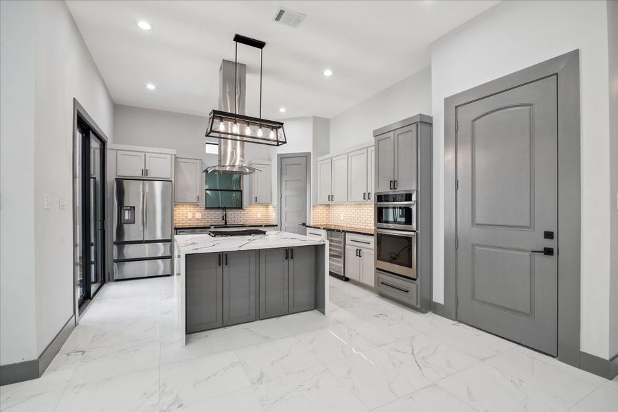 Custom cabinetry pairs seamlessly with a large waterfall island and stainless steel appliances. Sleek vent hood and statement lighting create a strong focal point, while the subway tile backsplash and under-cabinet lighting bring in timeless detail.