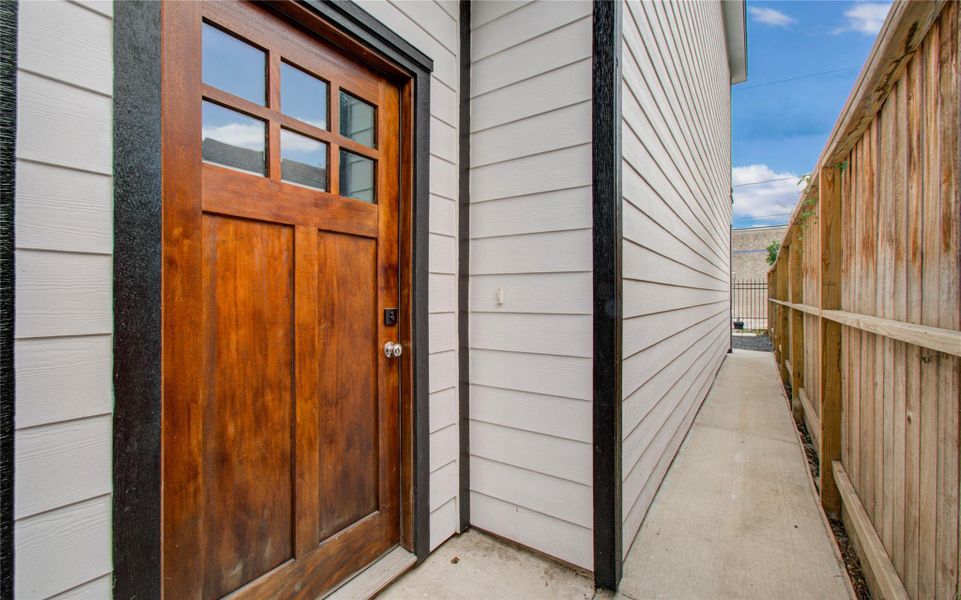 Side entrance to the home with a wooden door featuring glass panels. It opens onto a walkway bordered by a wooden fence, leading to an outdoor area. The siding is a bright white with black trim, offering a clean and modern look.
