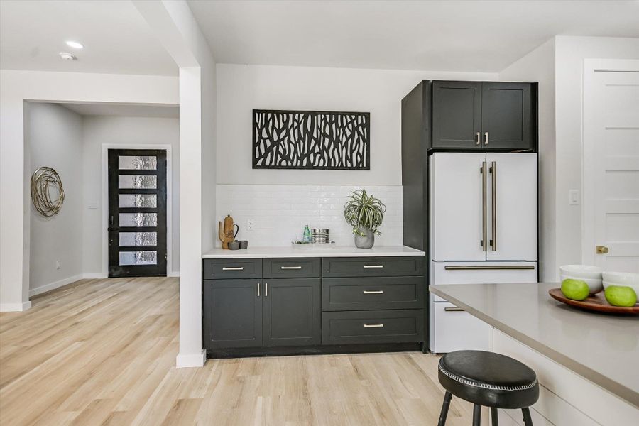 A well-lit kitchen with sleek dark cabinetry, modern appliances, and light hardwood flooring extends towards the entry.