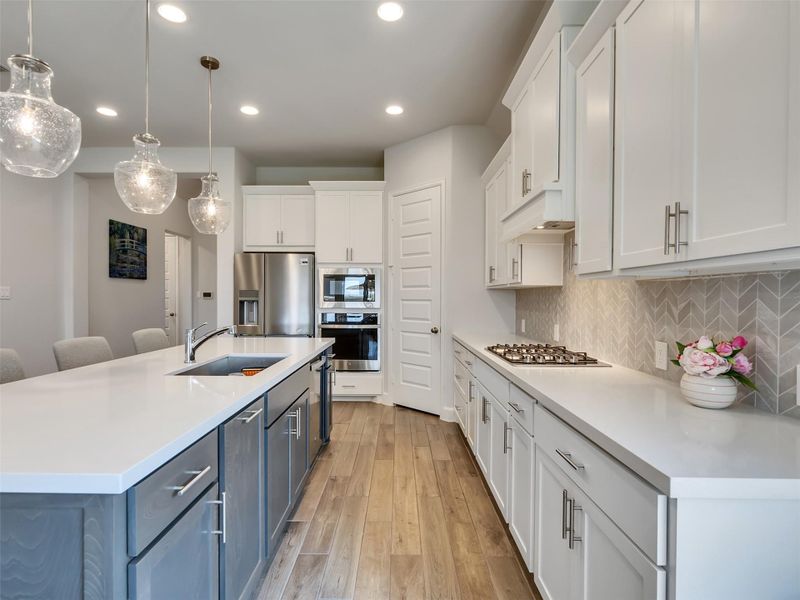 Elegant kitchen featuring modern pendant lighting, sleek white cabinetry, and a spacious island with a sink. Stainless steel appliances and a stylish herringbone backsplash complete the contemporary look.