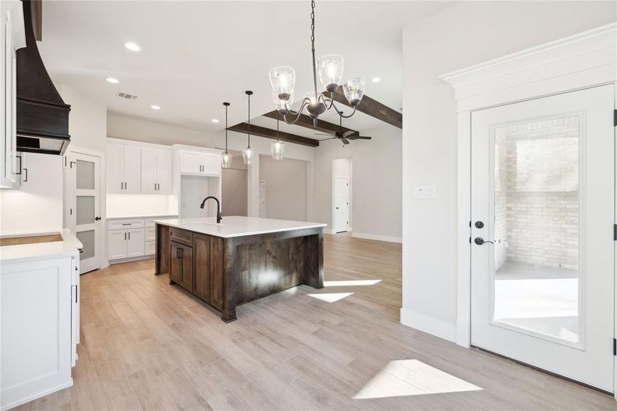 Kitchen with beamed ceiling, light hardwood / wood-style floors, a kitchen island with sink, white cabinets, and ceiling fan with notable chandelier