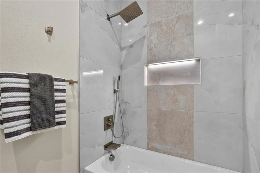 Bathtub with a large format tile surround featuring a rainfall showerhead, handheld shower, and an illuminated recessed niche