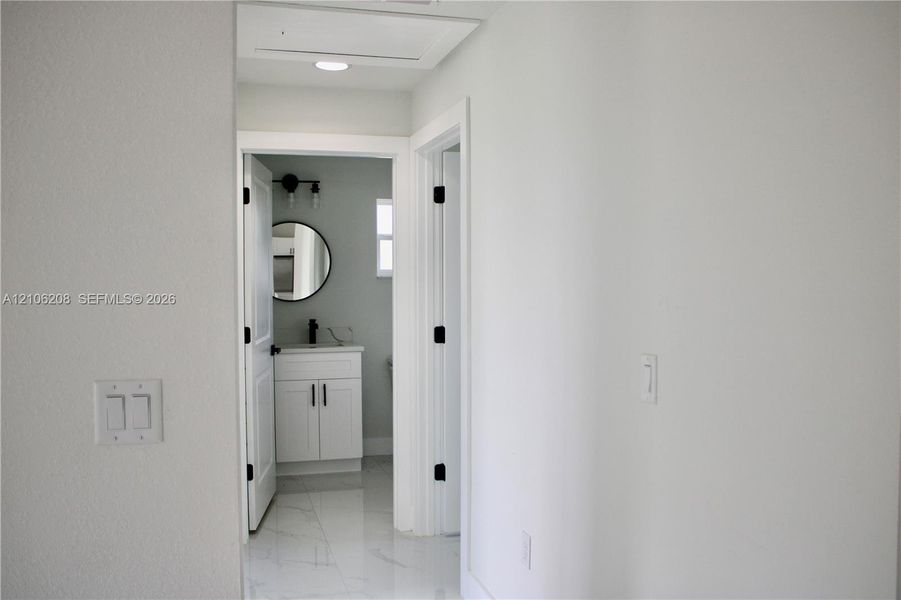 Hallway featuring light-colored tile flooring and recessed ceiling lighting, revealing a bathroom with a white vanity, matte black hardware, and a circular mirror
