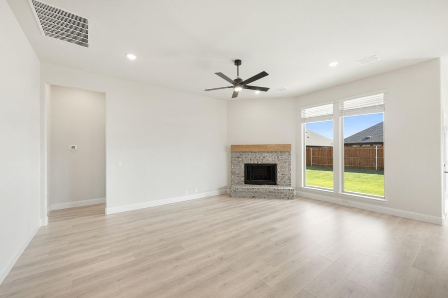 Open living room with light wood floors, brick fireplace, ceiling fan, and large window overlooking backyard