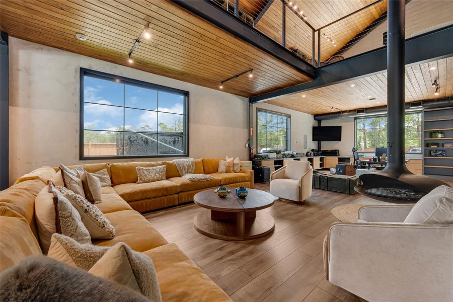 Open-concept design with a high wood-paneled ceiling and large windows for natural light. It includes a cozy sectional sofa, a contemporary fireplace, and sleek furnishings, creating a warm and inviting atmosphere. Perfect for relaxation and entertaining.