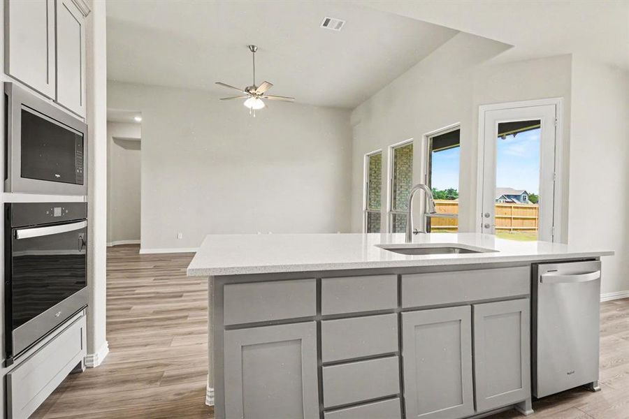 Open-concept kitchen featuring an island with a built-in sink and stainless steel dishwasher, light gray cabinetry, wood-finish flooring, and integrated stainless steel wall oven and microwave Open-concept kitchen featuring an island with a built-in sink and stainless steel dishwasher, light gray cabinetry, wood-finish flooring, and integrated stainless steel wall oven and microwave