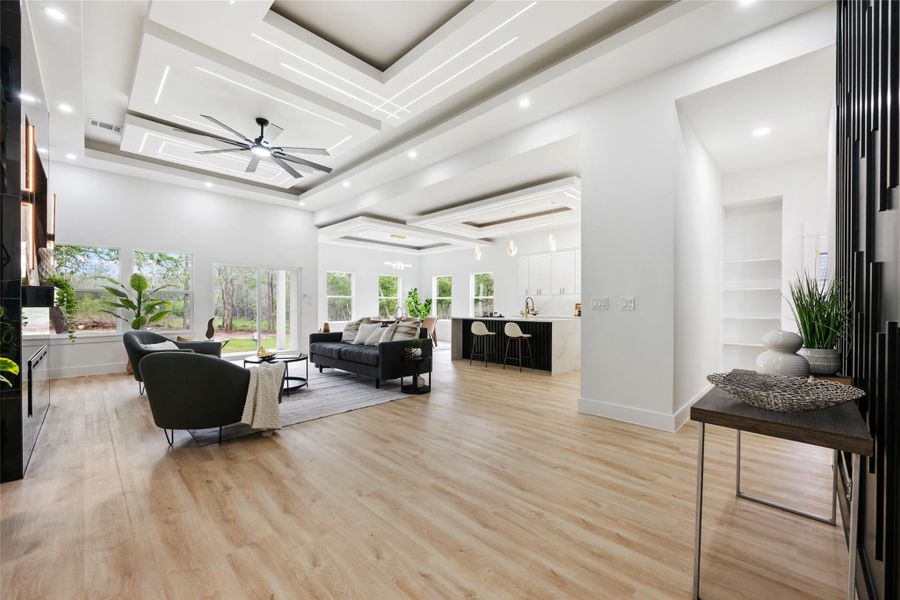 Expansive open-concept living area featuring light wood-finish flooring, a tiered tray ceiling with linear lighting and recessed lights, and a ceiling fan