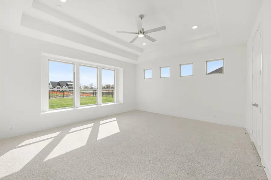 Empty bedroom with tray ceiling, ceiling fan, carpet flooring, and large windows overlooking a backyard Empty bedroom with tray ceiling, ceiling fan, carpet flooring, and large windows overlooking a backyard