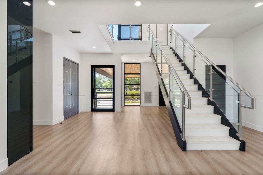 The staircase features floating light-toned treads on a black steel stringer, rising alongside a chrome and glass panel railing. The glass-paneled front entry door is visible below and the elevator landing sits just adjacent — form and function meeting at the entry of the home.