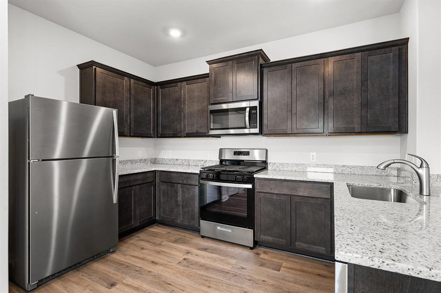 Kitchen featuring appliances with stainless steel finishes, light wood-type flooring, dark brown cabinetry, and light stone countertops