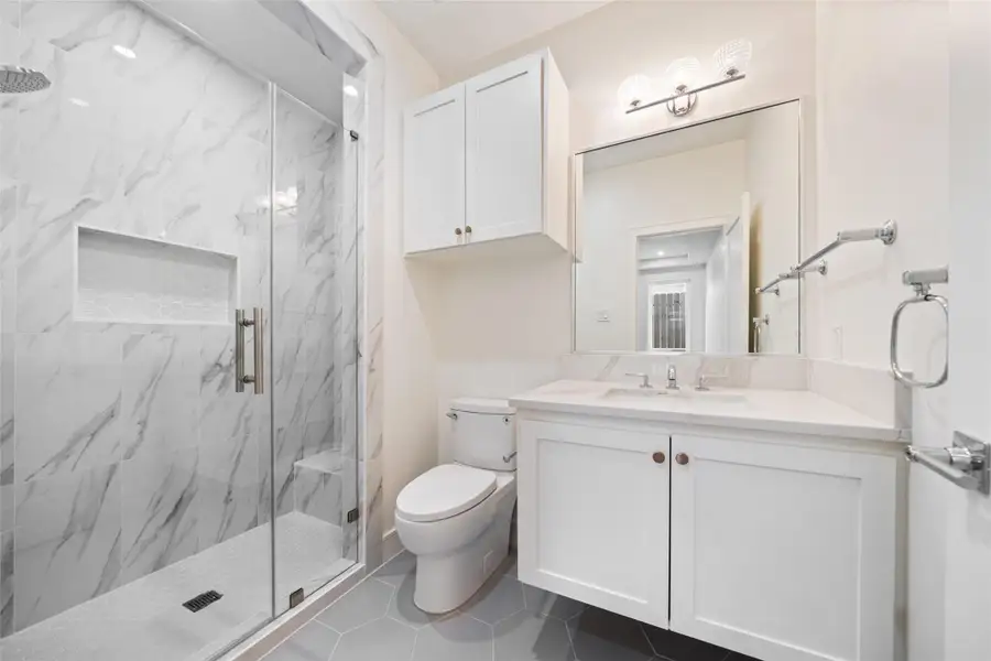 First floor en-suite bathroom features a sleek glass-enclosed shower with marble-style tiles, a spacious vanity with ample storage, and elegant fixtures. The neutral color palette and large mirror enhance the sense of space and light.
