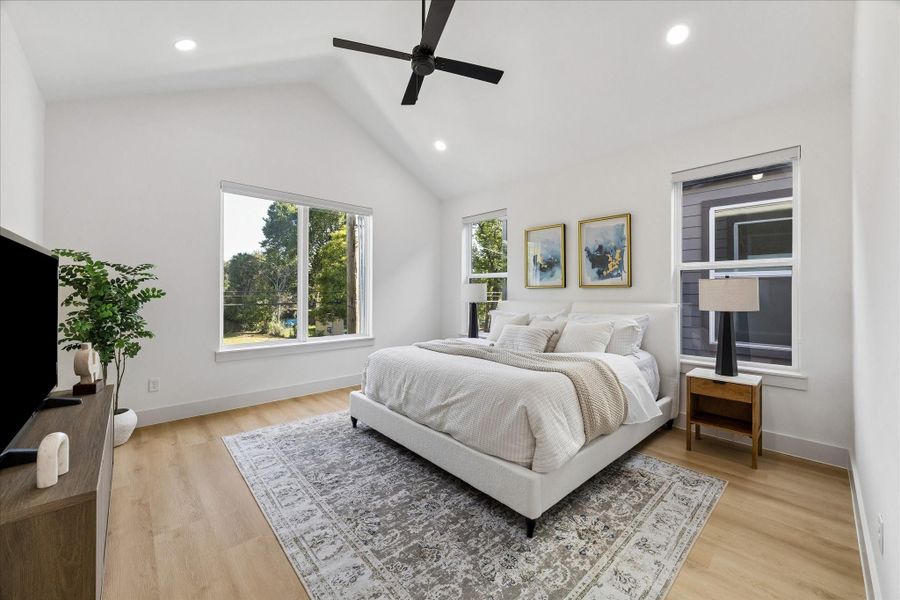Vaulted ceilings and oversized windows create a bright, airy retreat in the spacious primary suite. Warm wide plank flooring and a calming neutral palette enhance the room’s serene atmosphere, while the generous layout easily accommodates large furnishings with space to spare. Thoughtfully positioned windows invite abundant natural light throughout the day, making this an inviting space to relax and unwind.