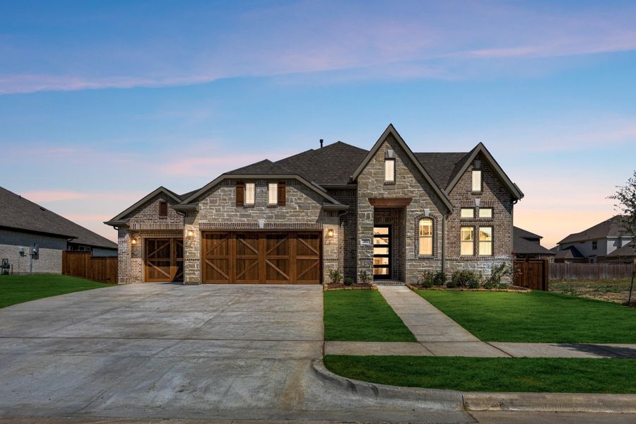 Single-story stone and brick home with three-car garage, wood doors, and manicured front lawn at dusk. Single-story stone and brick home with three-car garage, wood doors, and manicured front lawn at dusk.