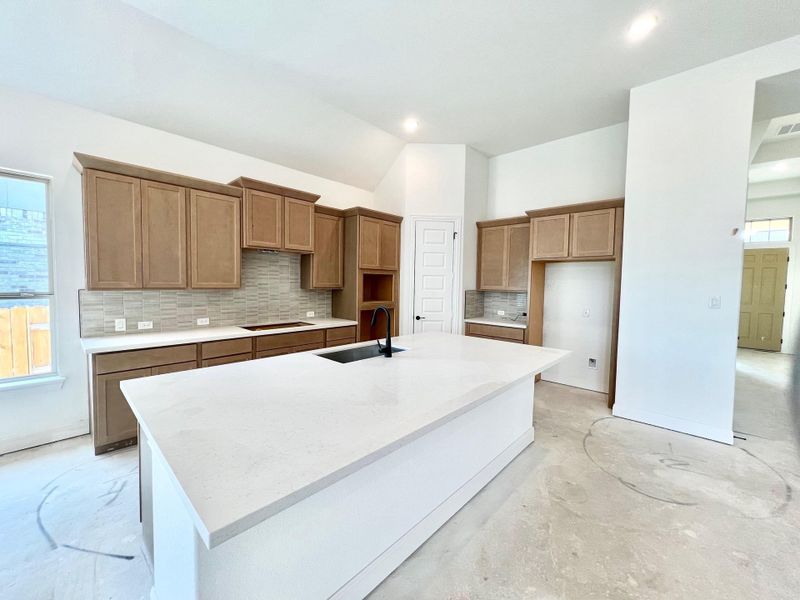 Kitchen with backsplash, light stone counters, a center island with sink, vaulted ceiling, and concrete flooring Kitchen with backsplash, light stone counters, a center island with sink, vaulted ceiling, and concrete flooring