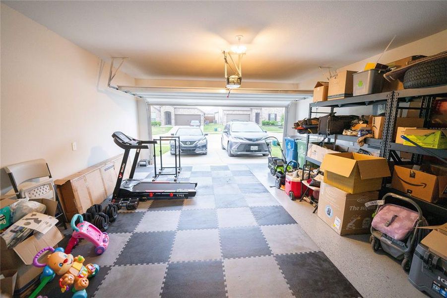 Spacious two-car garage with a checkered floor and overhead lighting, featuring built-in shelving and an automatic garage door opener Spacious two-car garage with a checkered floor and overhead lighting, featuring built-in shelving and an automatic garage door opener