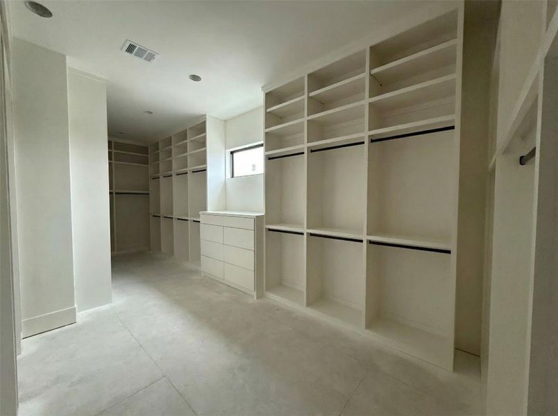 Primary Closet- Expansive walk-in closet with floor-to-ceiling custom built-ins, dual hanging sections, custom drawers, and laundry room basket pass through and beautiful natural window light. (Construction Photo as of 3/8/26 - Edited)