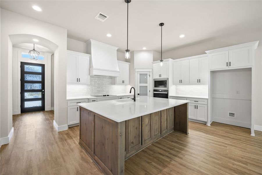 Kitchen featuring two tone cabinets, a center island with sink, pendant lighting, tasteful backsplash, and dark wood finished floors