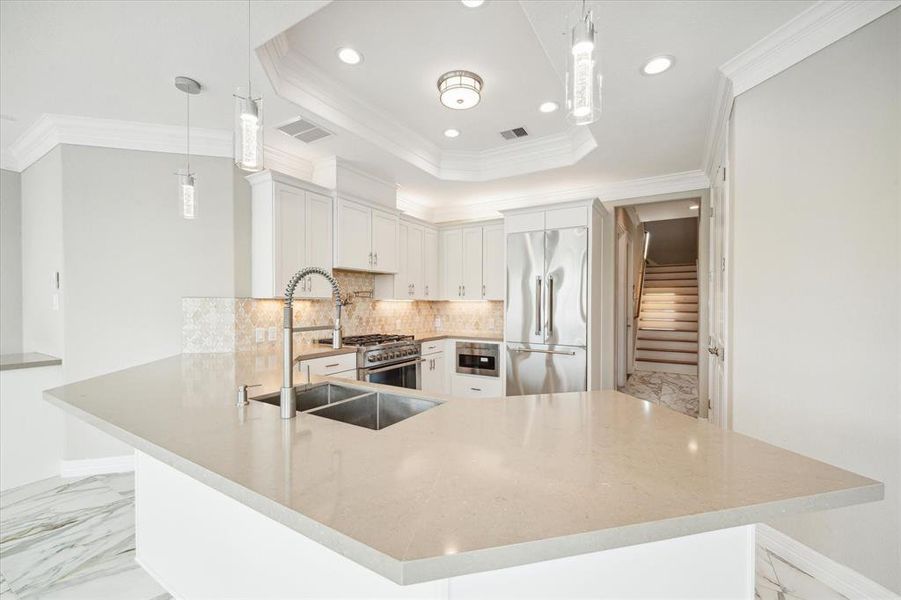 This is a modern, well-lit kitchen featuring quartz countertops,white cabinetry, top quality stainless steel appliances, a large island with a sink, and elegant pendant lights. There's also a decorative backsplash and a view of the waterway.