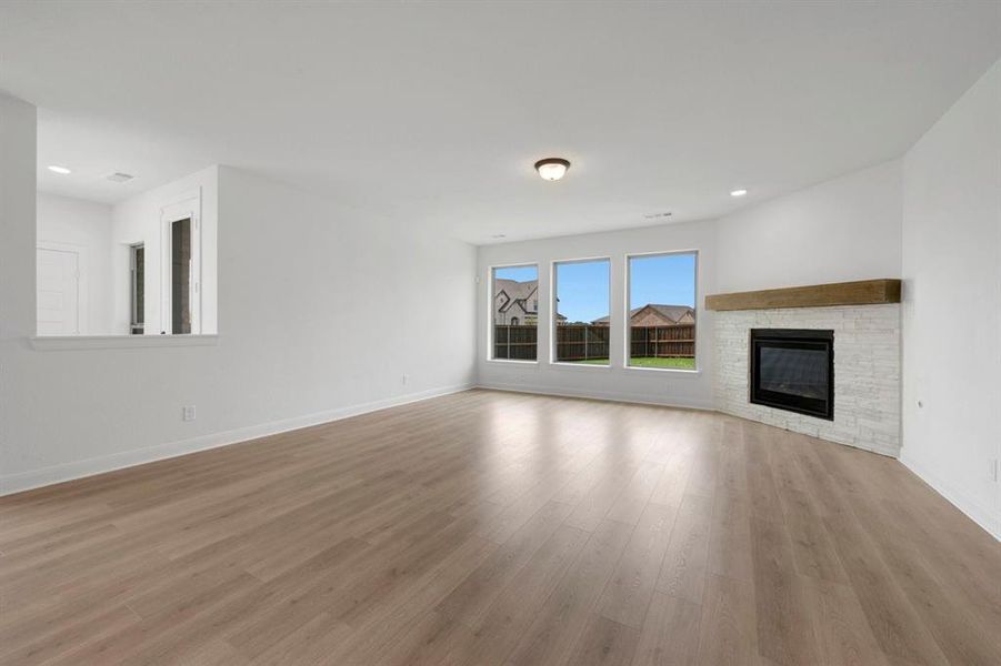 Spacious living area featuring wood-finish flooring, a white-stacked stone fireplace with a substantial wood mantel, and a trio of large windows