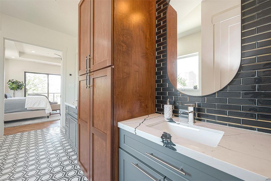 This is a modern bathroom featuring a sleek vanity with gray cabinetry and wood accents, a unique black tile backsplash, and an elegant oval mirror. Geometric patterned floor tiles add a stylish touch, and the room opens into a bedroom, suggesting an en suite layout. This is a modern bathroom featuring a sleek vanity with gray cabinetry and wood accents, a unique black tile backsplash, and an elegant oval mirror. Geometric patterned floor tiles add a stylish touch, and the room opens into a bedroom, suggesting an en suite layout.