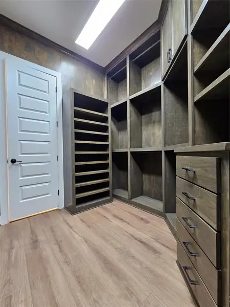 This photo shows a spacious walk-in closet with custom wood shelving and drawers, ample storage space, and a modern design. The room is well-lit, featuring a light wood floor and a sleek white door.