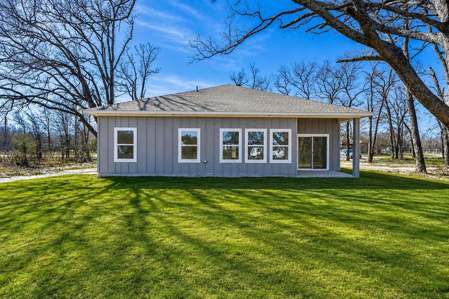 Rear exterior featuring grey vertical siding, a shingled roof, multiple windows with white trim, a sliding glass door, and an attached concrete patio Rear exterior featuring grey vertical siding, a shingled roof, multiple windows with white trim, a sliding glass door, and an attached concrete patio