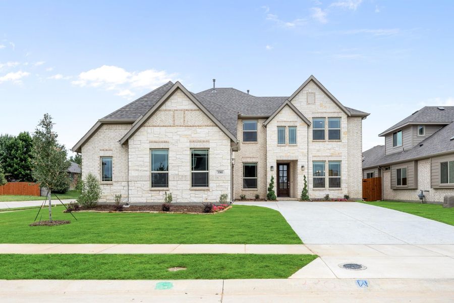 Two-story home with light stone exterior, dark roof, and concrete driveway with green front lawn