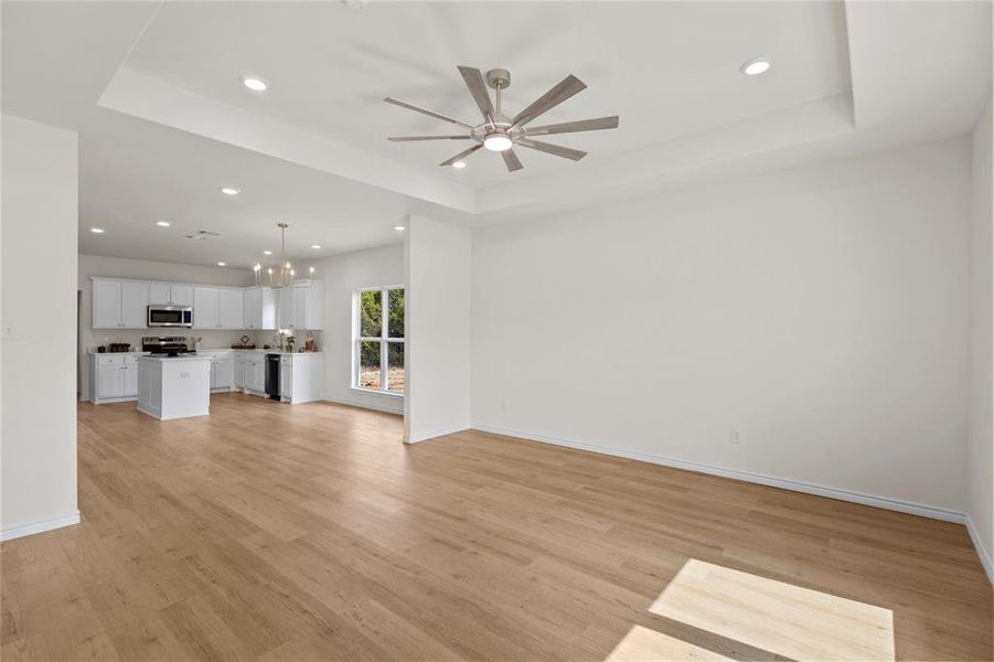 Unfurnished living room featuring a tray ceiling, recessed lighting, light wood-type flooring, a chandelier, and ceiling fan Unfurnished living room featuring a tray ceiling, recessed lighting, light wood-type flooring, a chandelier, and ceiling fan
