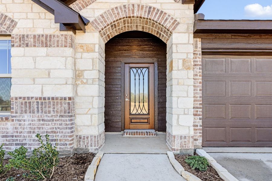 Front entry with stone arch, wood door with iron glass panel, and brick exterior with garage door visible