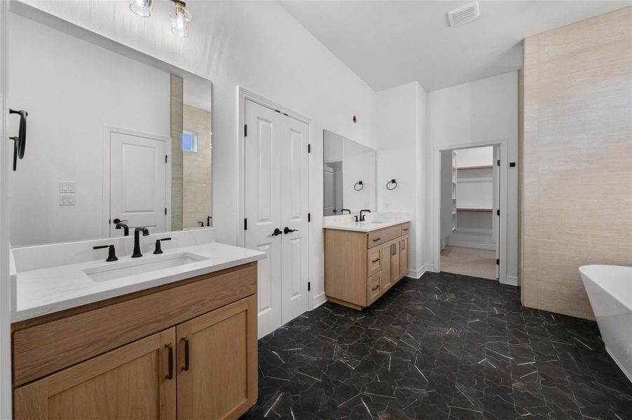 Black patterned tile flooring anchors this bright bathroom, featuring dual vanities and an adjacent walk-in closet. Black patterned tile flooring anchors this bright bathroom, featuring dual vanities and an adjacent walk-in closet.