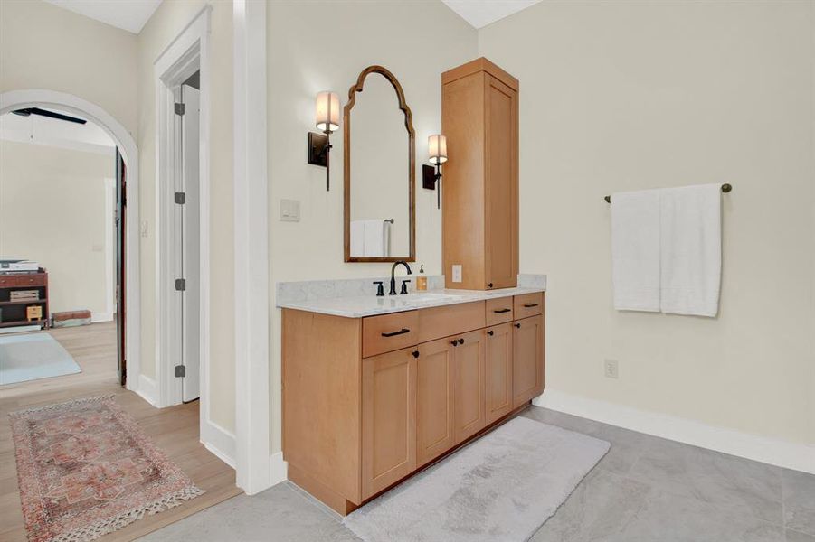 Beautiful wooden vanities with ample storage, marble countertops, are complemented by a large mirror and warm lighting. The room has a neutral color scheme and includes elegant fixtures, with a glimpse of an adjoining area through an arched doorway.