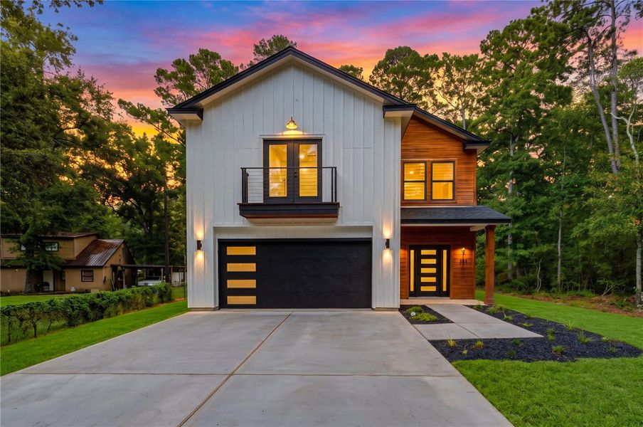 Stunning modern home with a sleek façade, featuring a contrasting dark garage door and elegant wood accents. The expansive driveway and well-lit exterior enhance its contemporary charm, set against a backdrop of lush trees and a vibrant sunset.