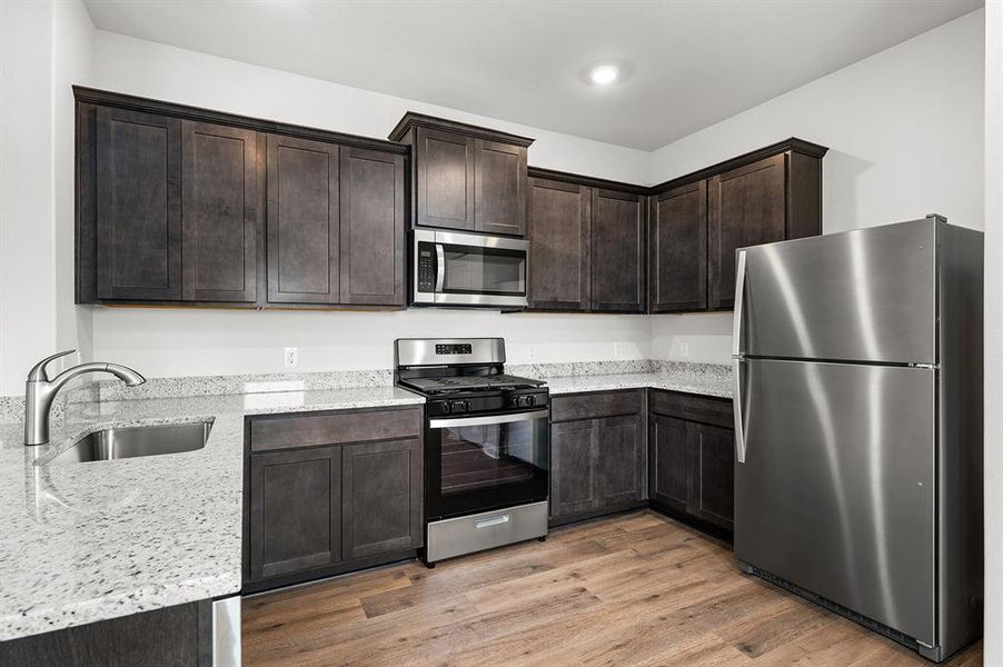 Kitchen featuring stainless steel appliances, light wood-style flooring, dark brown cabinets, and light stone countertops Kitchen featuring stainless steel appliances, light wood-style flooring, dark brown cabinets, and light stone countertops