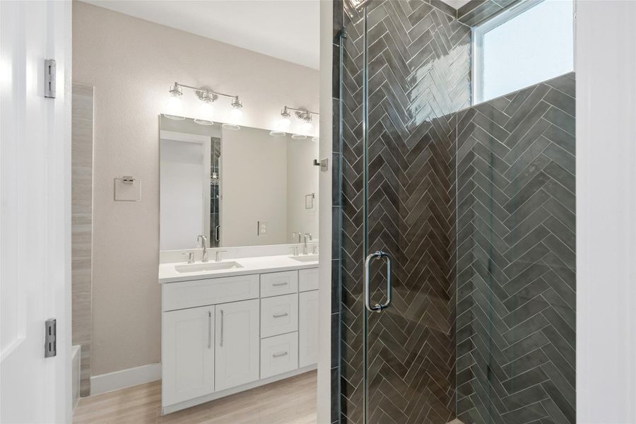 This bathroom features a modern design with a sleek double vanity, ample storage, and stylish lighting. The glass-enclosed shower showcases elegant herringbone tile work, complemented by a frosted window for natural light.