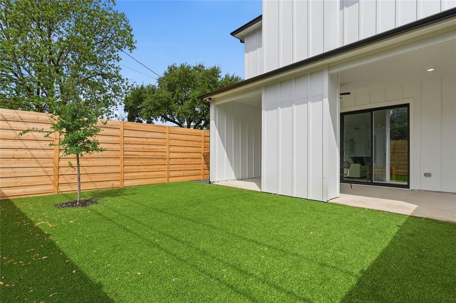 Expansive outdoor living area featuring a covered patio with recessed lighting, artificial turf, a newly installed horizontal plank privacy fence, and modern white board and batten siding