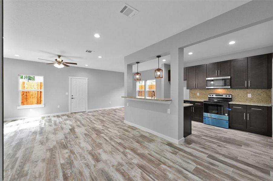 Discover contemporary elegance in this open-concept living area, boasting a sleek kitchen with rich dark cabinets and high-end stainless appliances, an inviting breakfast bar, and a sunlit living space with stylish wood-inspired flooring.